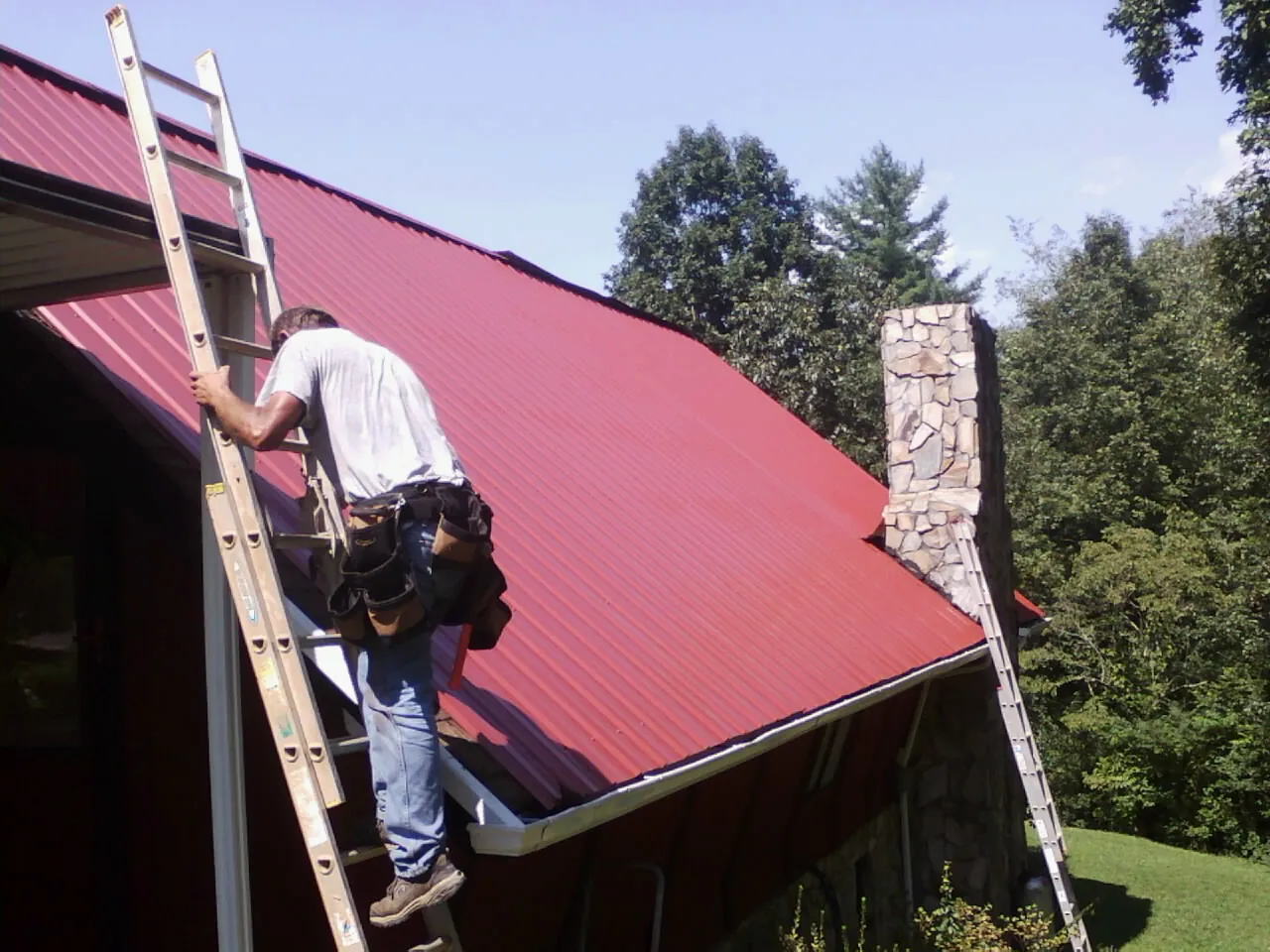 Roofer on a ladder beside a red metal roof and stone chimney — Soffit & Fascia Repair by Roofing Brothers in Central Valley